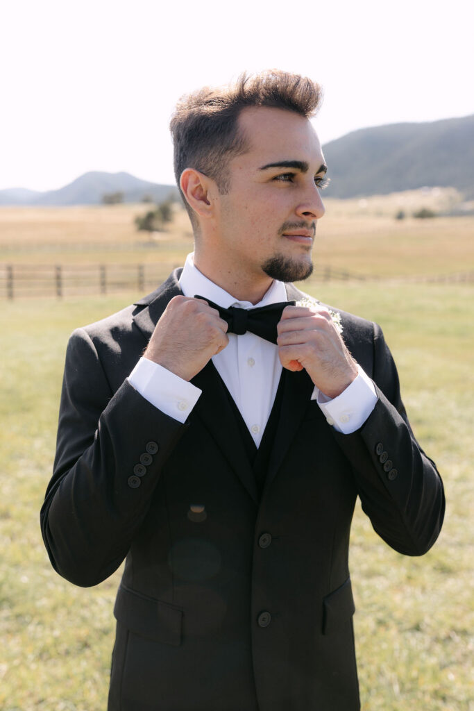 Groom adjusting his bow tie in front of the mountains at Spruce Mountain Ranch.