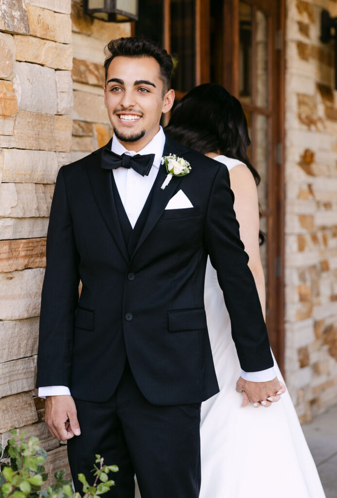Groom smiling in a black tuxedo outside the rustic lodge at Spruce Mountain Ranch during their first touch.
