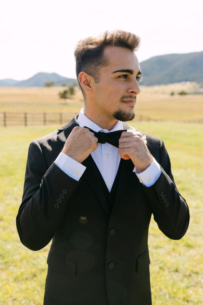Groom adjusting his bow tie in front of the mountains at Spruce Mountain Ranch.