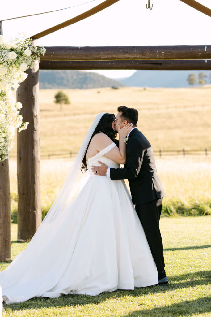 Bride and groom sharing first kiss under the wooden arch at Spruce Mountain Ranch.