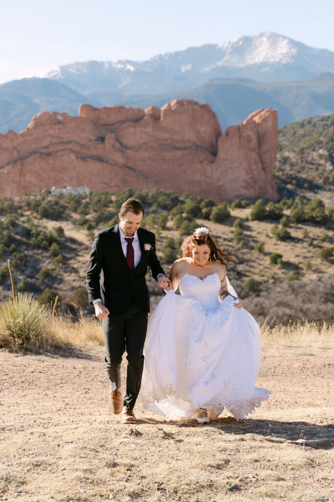 Candid wedding photography of couple running in front of red rock formations and Pikes Peak during their elopement in Garden of the gods in Colorado