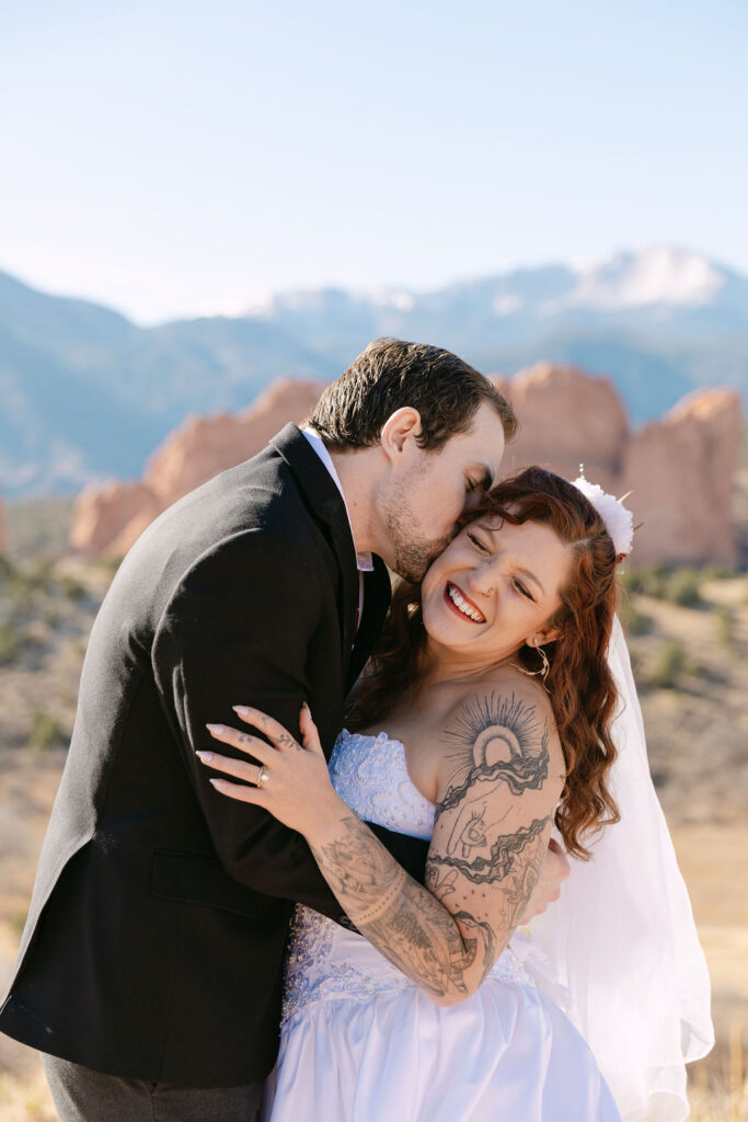 Natural, candid photo of groom kissing bride on the cheek as they hug in front of Mesa Overlook during their Colorado springs elopement in garden of the gods