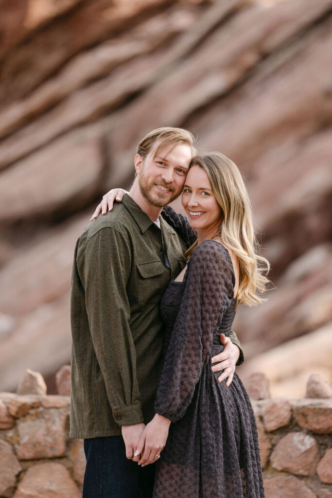 Classic engagement photography with couple hugging and smiling at the camera in front of a red sandstone formation in Colorado 