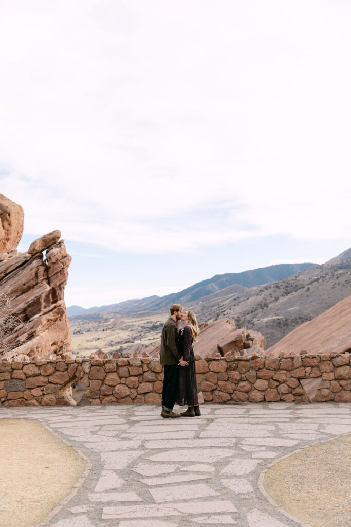 Far away scenery engagement photo of couple kissing with the Red Rocks Park in the background and blue sky