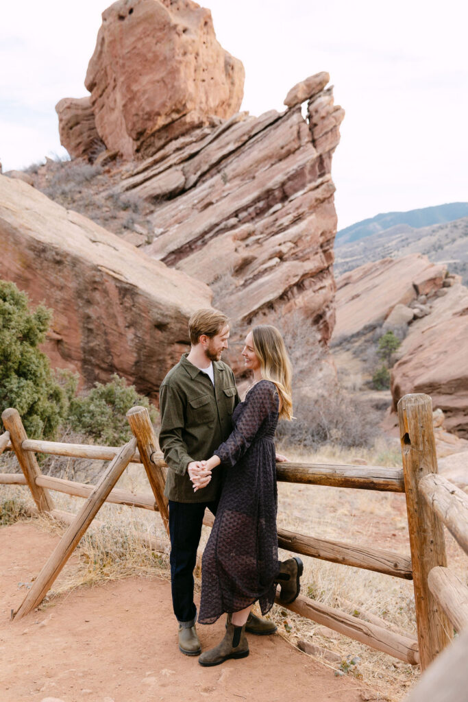 Candid engagement photos of couple holding hands leaning on a fence at red rocks park in morrison, Colorado 