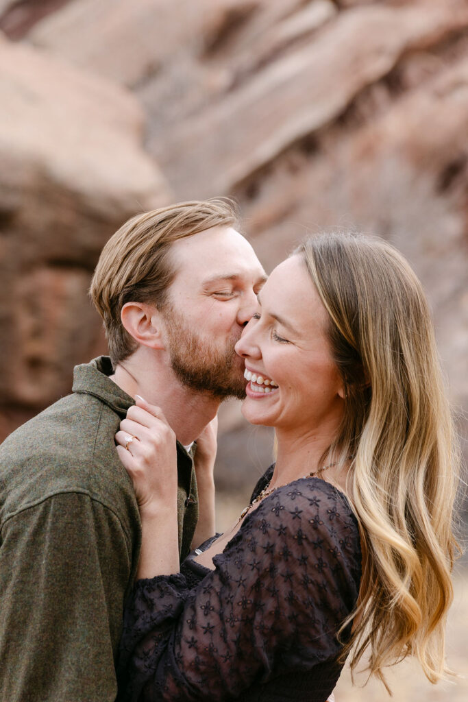 Candid, artful engagement photo of the man giving his fiancee rapid fire kisses as she holds onto his collar and laughs in front of the red sandstone rocks 