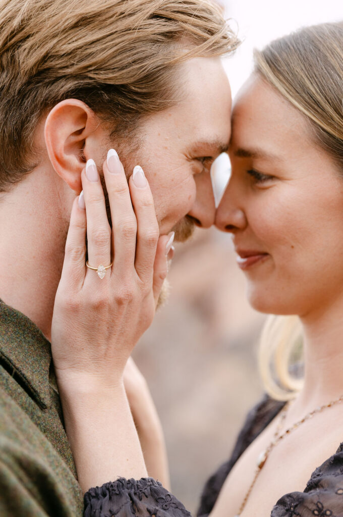 Romantic engagement photo in Colorado where the woman is embracing the man's cheek with her ring hand and they are nose to nose soft smiling in the background 