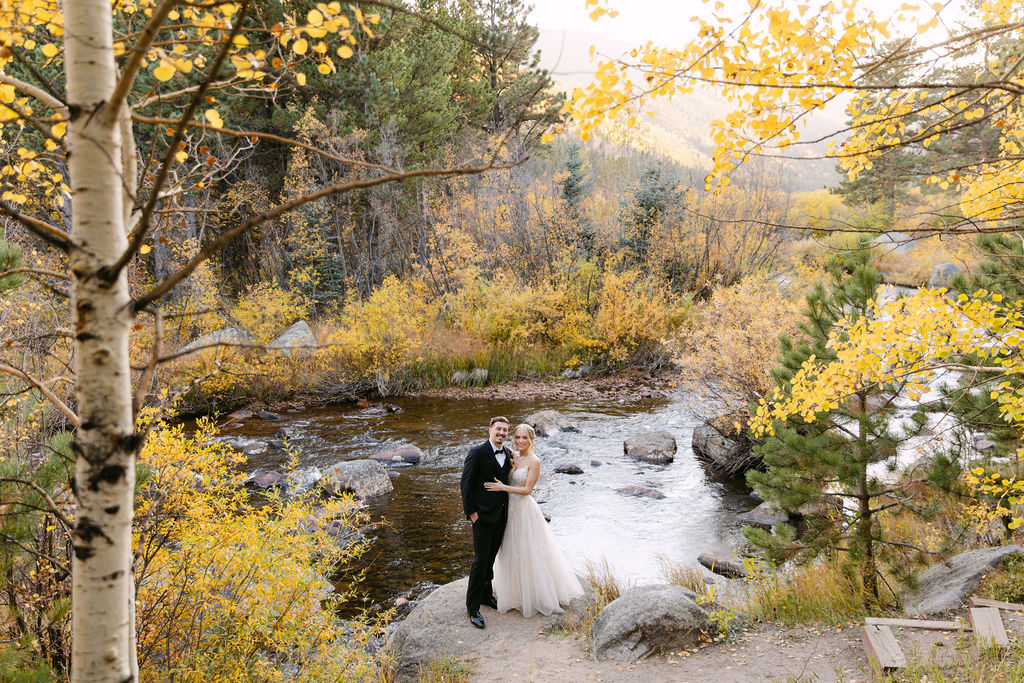 The couple smiles together during fall wedding portraits beside the river at Wild Basin Lodge in Allenspark, Colorado.