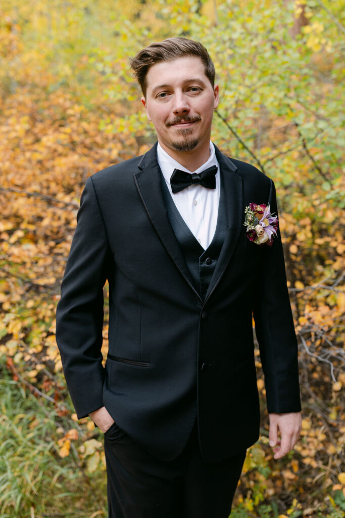A groom stands smiling in fall foliage  during his outdoor Colorado wedding. 
