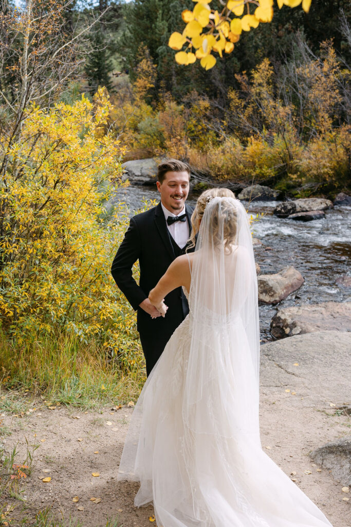 The couple holds hands by a river during their first look at their Colorado mountain wedding.