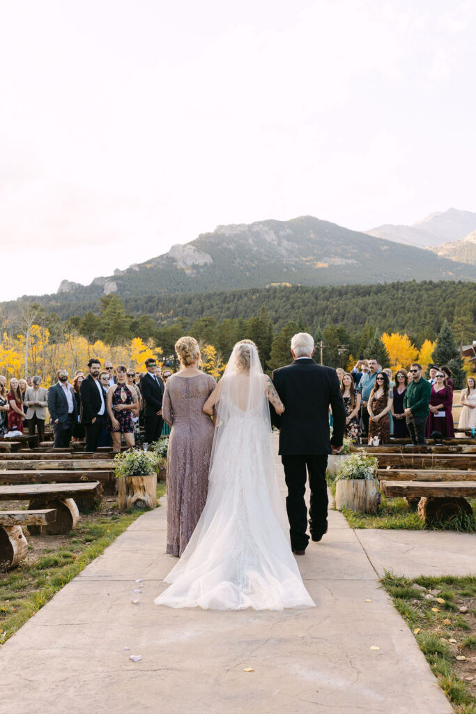 The bride walks down the ceremony aisle with guests seated outdoors at Wild Basin Lodge in Allenspark, Colorado, in Rocky Mountain National Park.