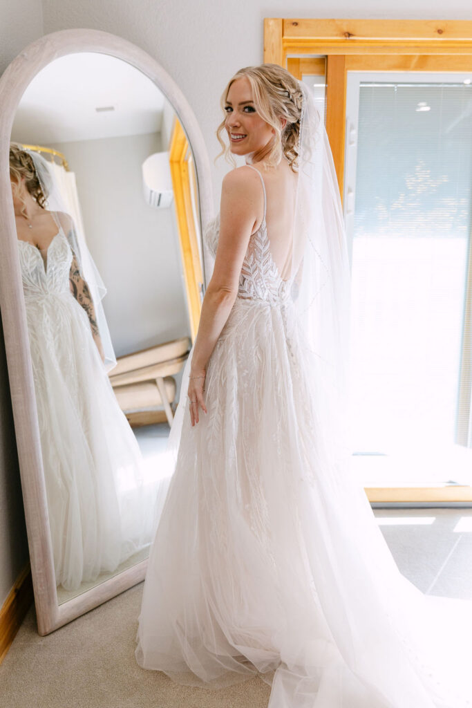 The bride shows the back of her wedding dress near a window at Wild Basin Lodge in Allenspark, Colorado, in Rocky Mountain National Park.