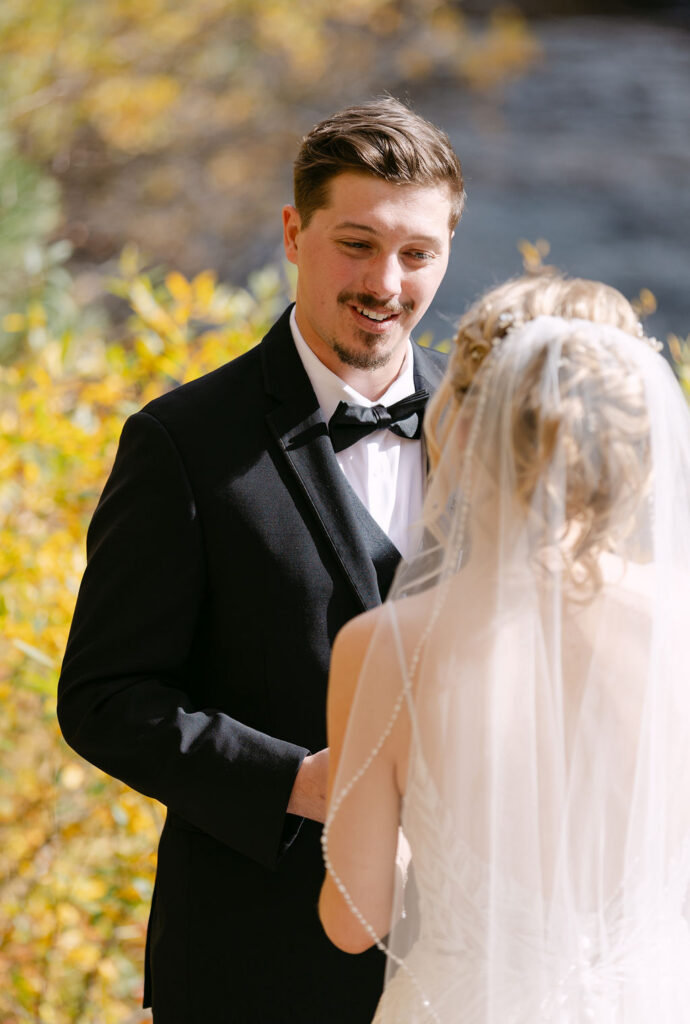 The groom smiles at the bride during their riverside first look at a mountain lodge wedding. 
