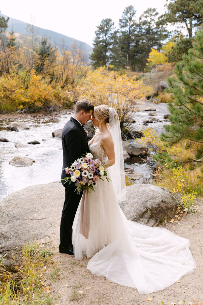The couple stands together holding a bouquet by the river at Wild Basin Lodge in Allenspark, Colorado, within Rocky Mountain National Park.