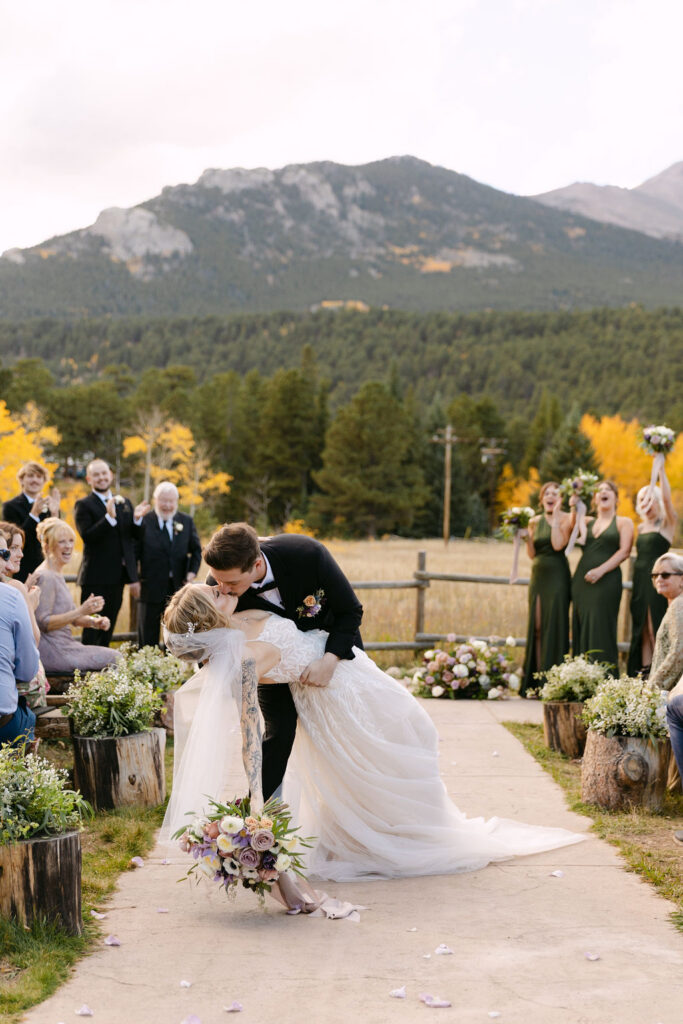 Bride and groom sharing a dip and kiss at the end of the aisle in a meadow with mountain views after their wedding ceremony. 